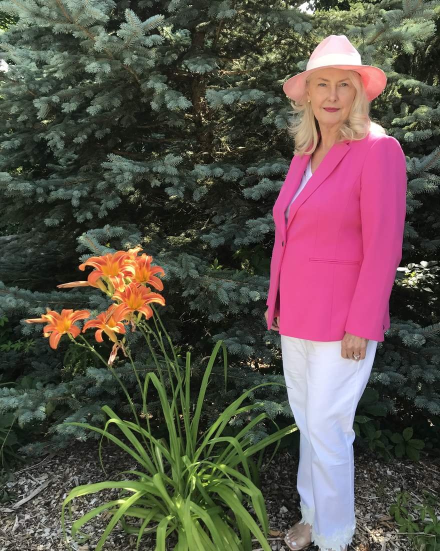 Wendy Baldwin standing beside orange daylilies in a garden, wearing a pink hat and jacket